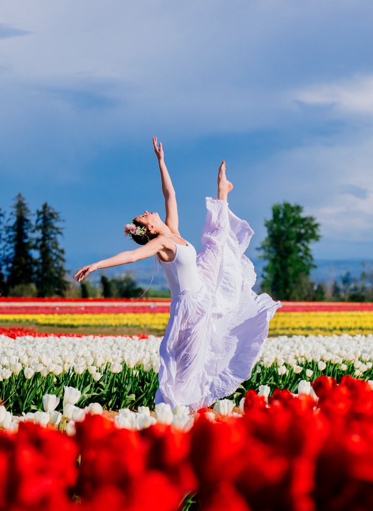 Ballet dancer wearing a white dress and flower crown dances gracefully in a colorful tulip field under a blue sky, USA.