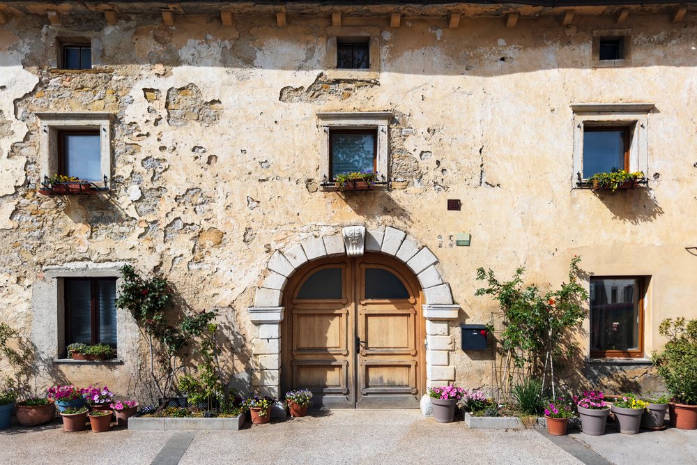 Weathered Building, featuring a stone-arched wooden door, climbing plants, and flower pots decorating the exterior walls, landscape, cityscape, Vipava, Slovenia.