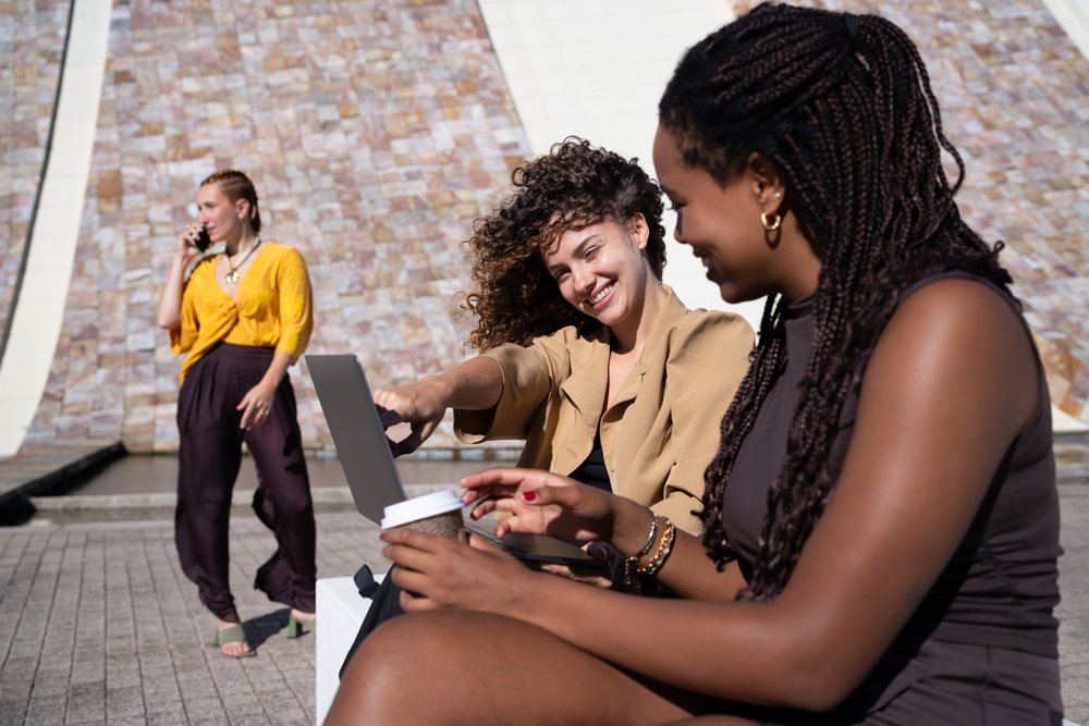 Three women engage outdoors on a sunny day, one using a laptop, another holding a coffee cup, and a third speaking on the phone, Spain.