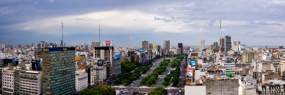 Panoramic cityscape of Buenos Aires featuring a busy avenue with vehicles, high-rise buildings, and a cloudy sky.