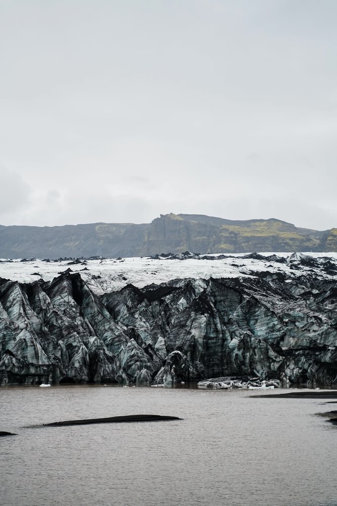 A glacier with icebergs floating in a lake in front of rocky cliffs under an overcast sky, Iceland.
