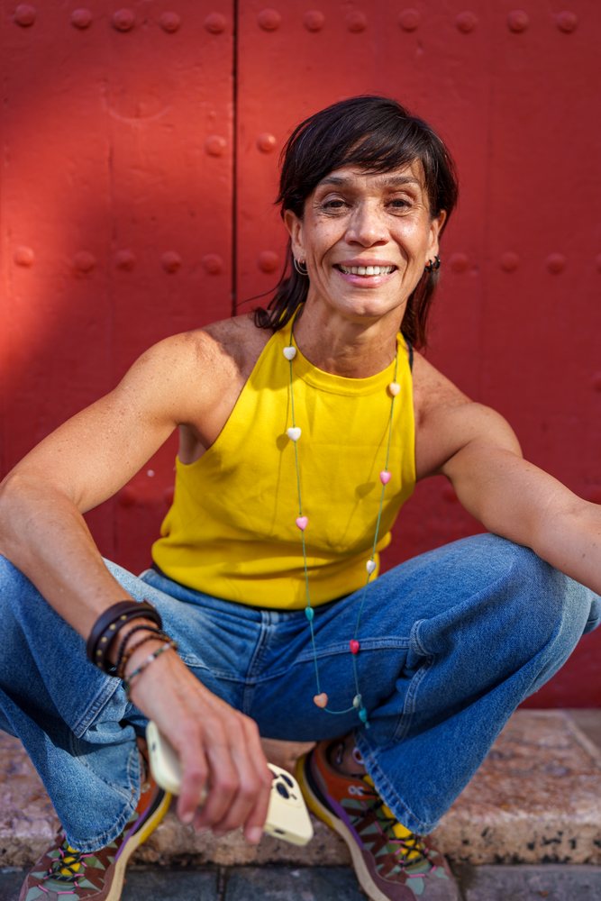 Woman sitting casually in front of red door wearing yellow sleeveless top and jeans outdoors, retro, vintage, Málaga, Spain.