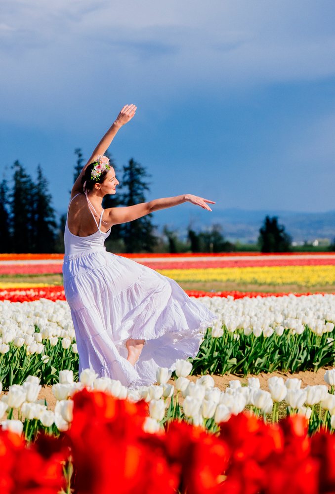 Female Ballet dancer in a white flowing dress and a floral crown dances gracefully amid colorful tulips outdoors, USA.