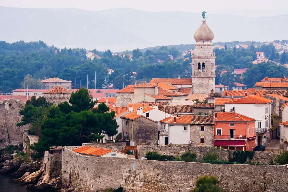 Old town of Krk Island with stone walls, red tiled roofs, and a church tower topped by a statue near the coast, Croatia.