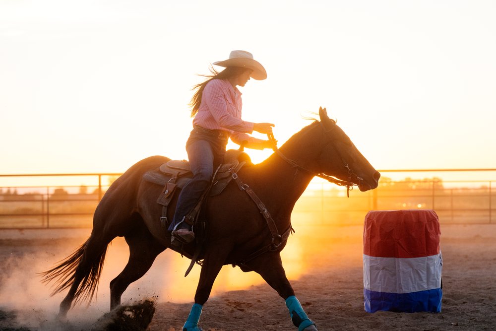 Female cowgirl rides a horse wearing saddle and reins near a red, white, and blue barrel in a dusty rode arena at sunset, Southwest USA.