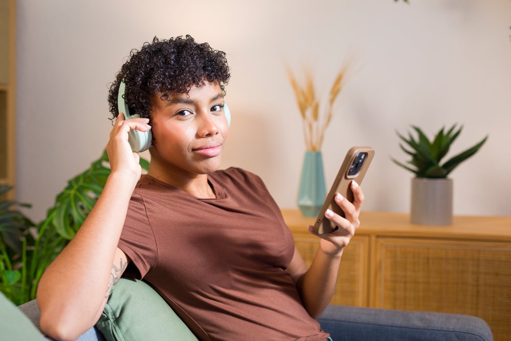 Young woman sitting indoors on a sofa wearing headphones and holding a smartphone, with tattoo visible and plants in the background.