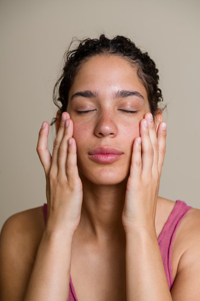 Woman with natural skin gently touching her face with eyes closed in a neutral setting during a beauty shoot, Spain.