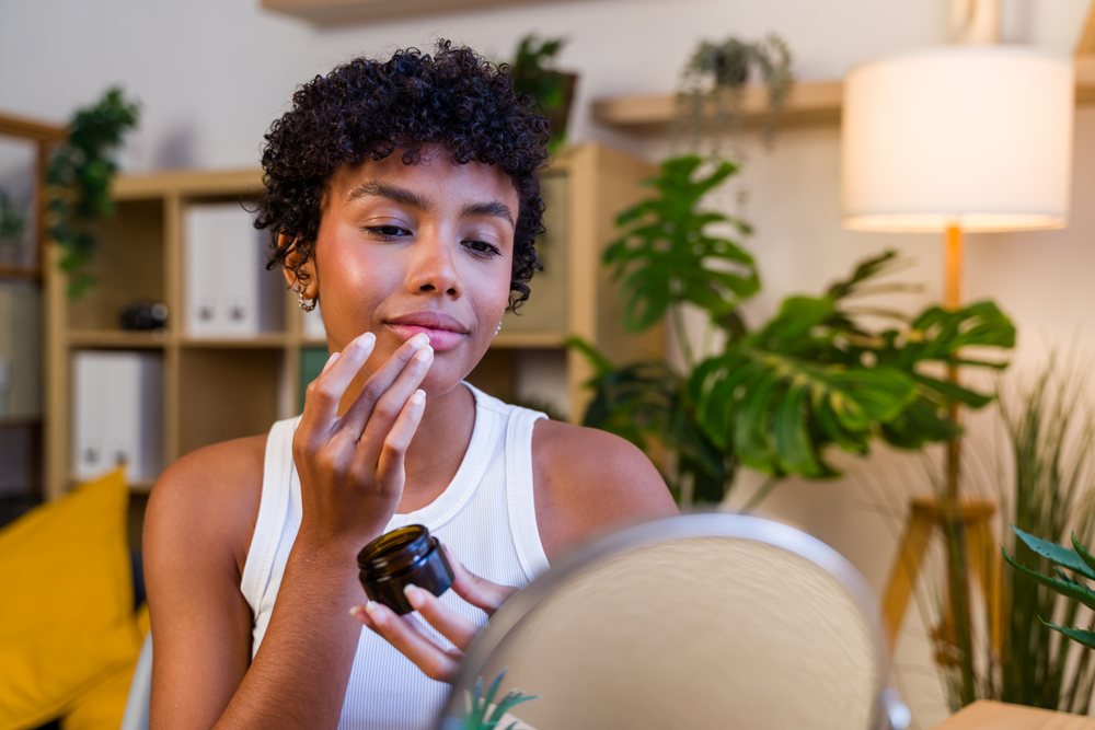 Woman applying face cream while looking in a mirror inside a living room with plants and a lamp, Spain.