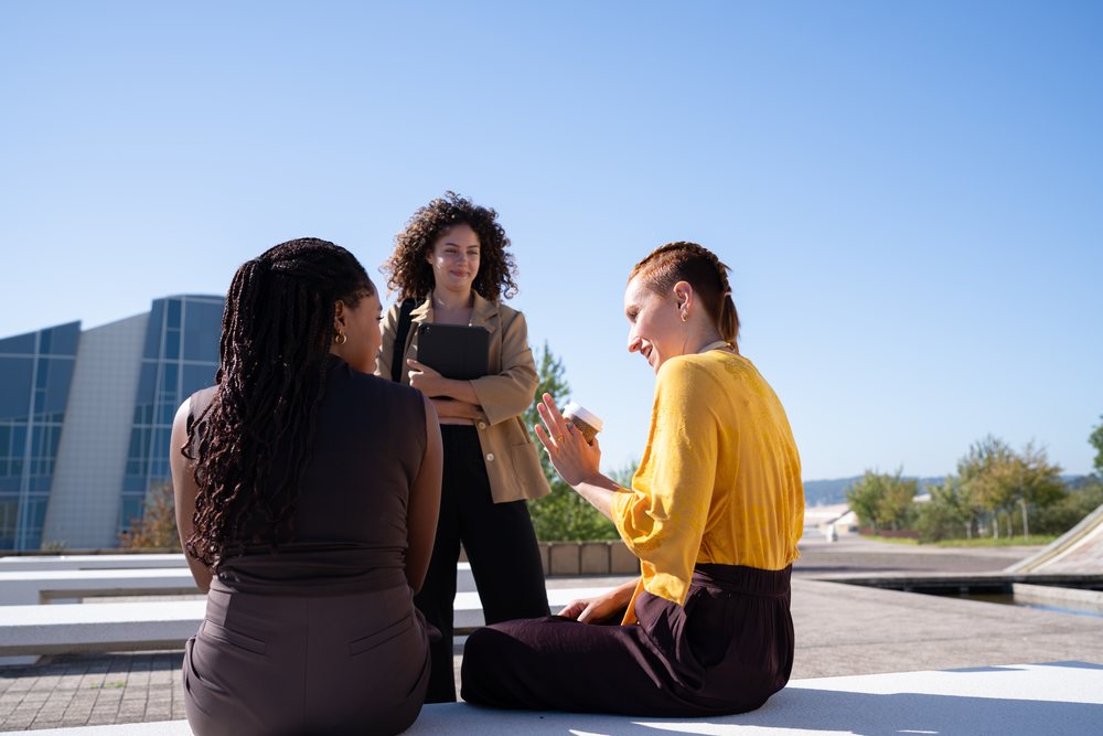 Three women engage in an outdoor conversation near modern glass buildings on a sunny day, Spain.