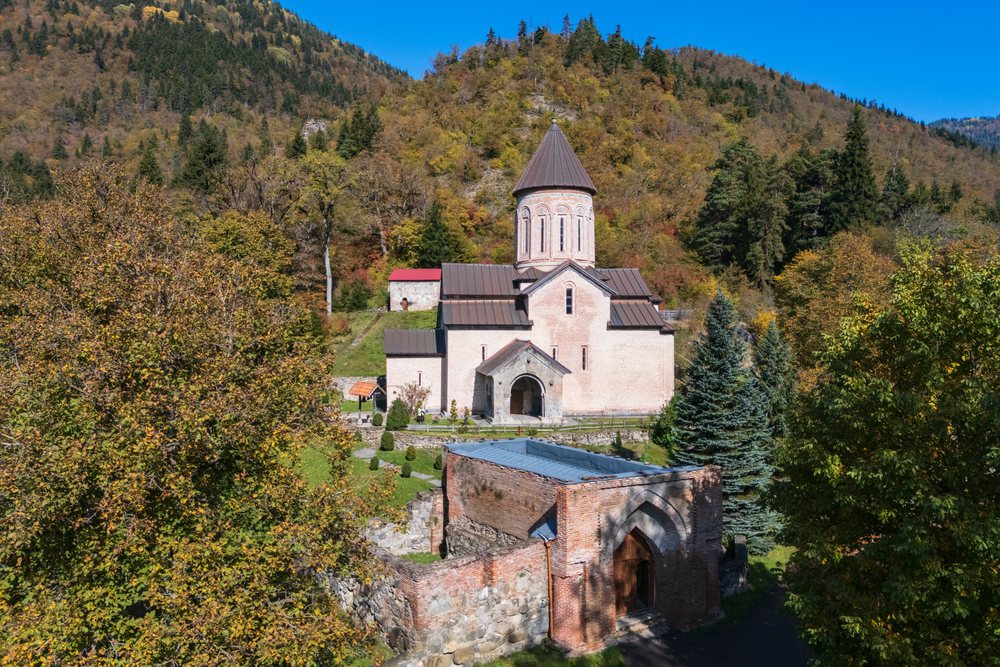 Tsagveri Timothesubvani Monastery with conical tower amid autumn trees and mountains under a clear blue sky, Georgia.