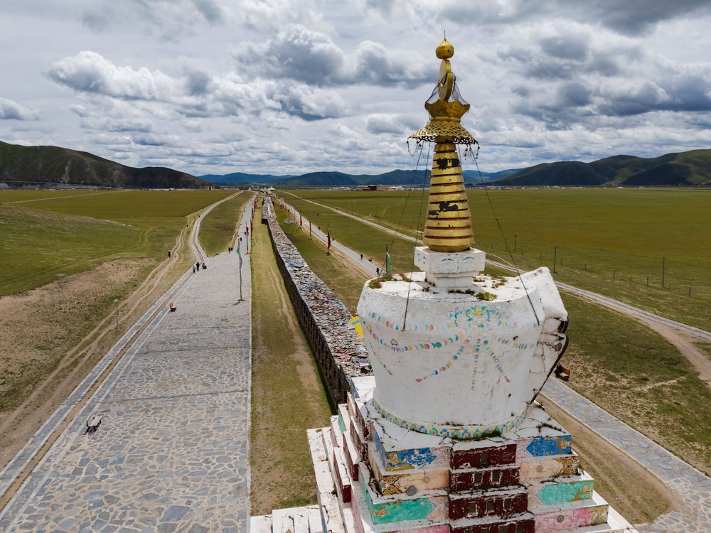The Bagomah Mani Wall with a decorated stupa and flag poles beside a stone walkway in Kham Tibet's grassy landscape under cloudy skies, Sichuan Province, People's Republic of China.