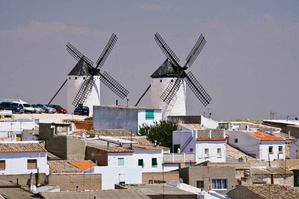 White windmills with black tops and sails tower above rooftops, Spain under a clear sky, La Mancha.