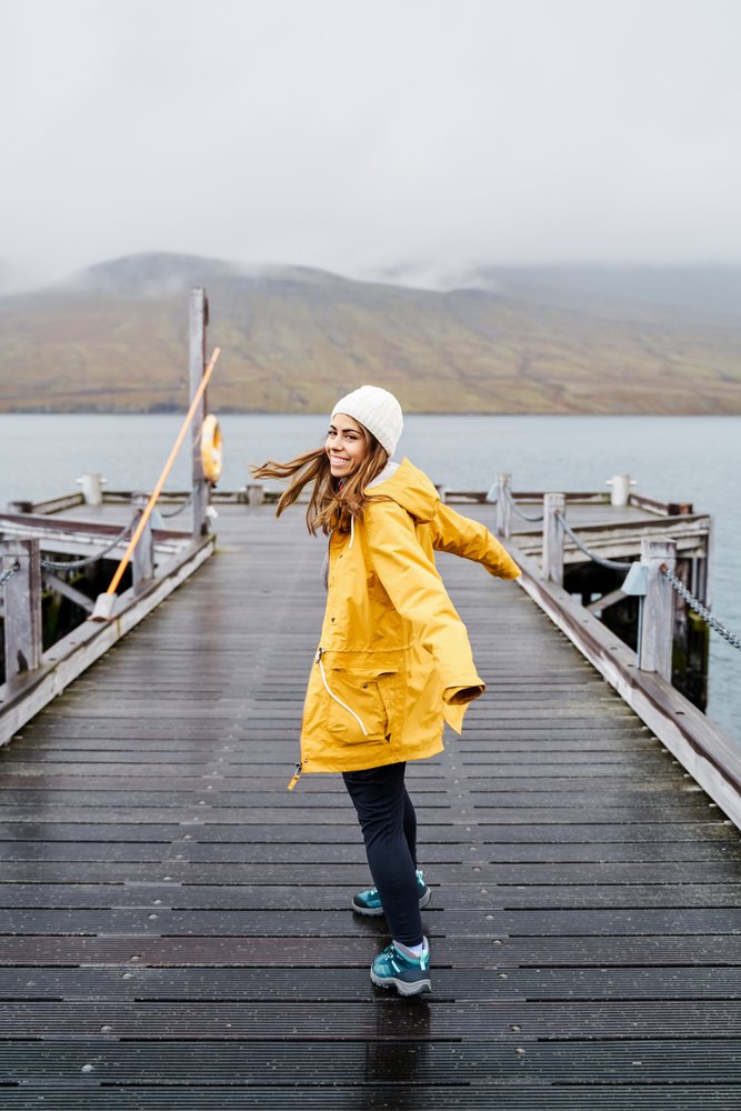 A woman wearing a yellow rain jacket and white beanie happily walks and spins on a wooden pier with foggy mountains and water in the background, Iceland.
