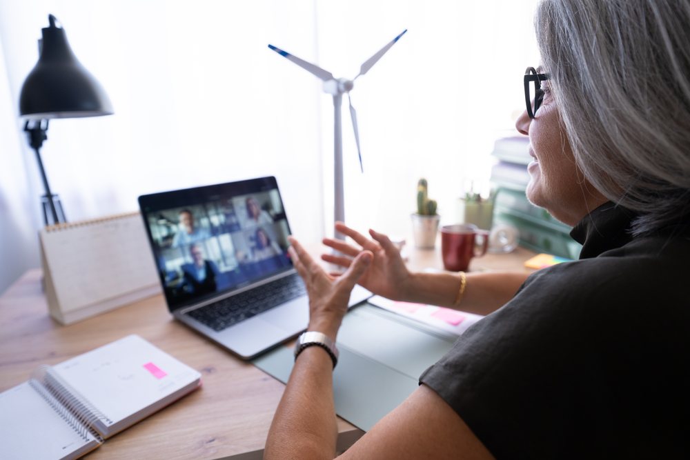 Gray-haired woman wearing glasses gestures while joining a video call at her home office desk with a laptop and office supplies, Spain.