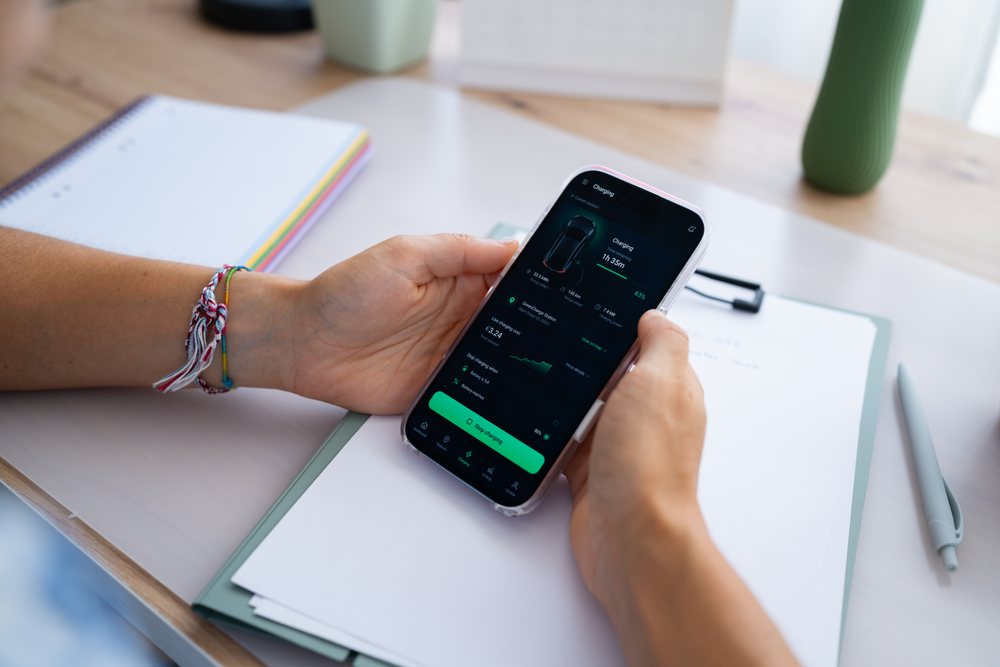 Hand holding smartphone displaying electric car charging app with charging status on wooden table beside notebook and pen, telecommuting, Spain.