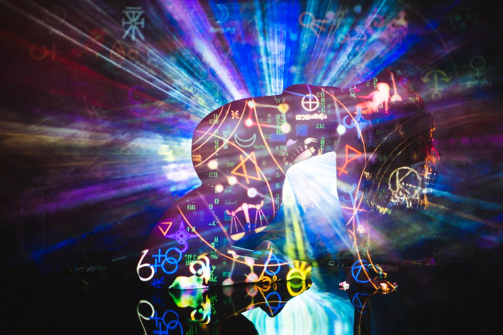 Nude person kneeling with multicolored symbolic projections and radiating light rays in an abstract artistic setting with dark background, pregnancy.