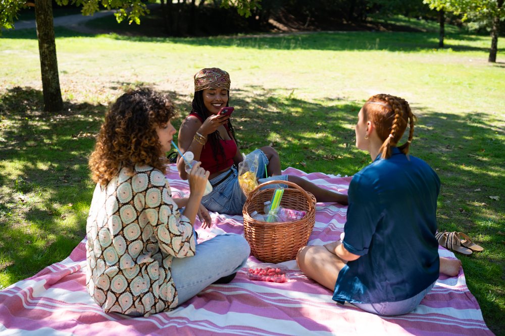 Three women sitting on a striped picnic blanket enjoying snacks and drinks in a sunny park under tree shade, Spain.