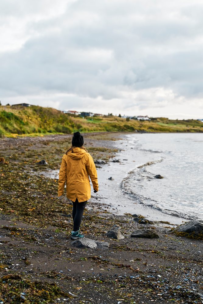 A white person walks on a rocky seashore under an overcast sky