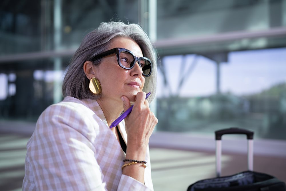 Businesswoman with gray hair and glasses holding a pen waiting with travel luggage in a modern airport terminal, Spain, Madrid.