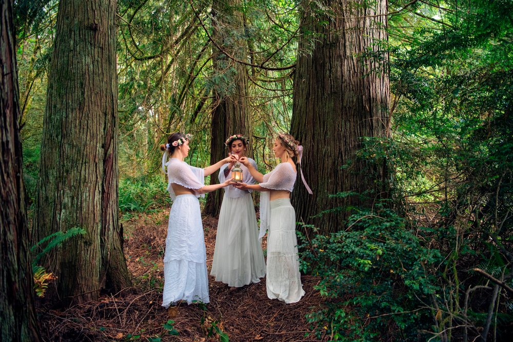 Three women wearing white dresses and flower crowns hold a lantern in a forest, muses, USA.