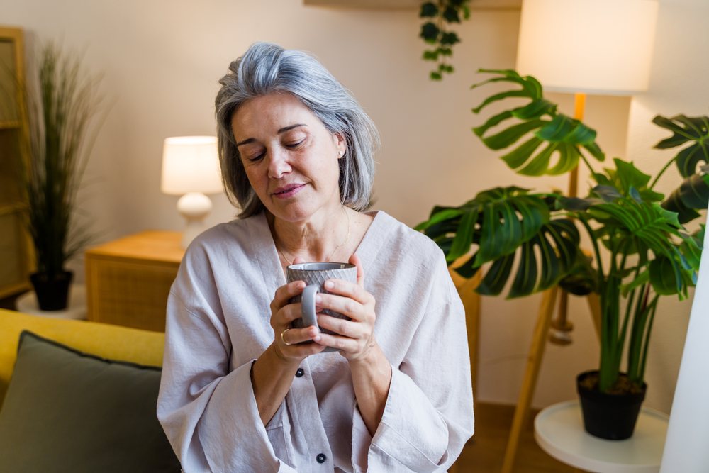 Woman with gray hair holding a cup and relaxing on a sofa in a cozy home setting with plants and warm lighting, Spain.