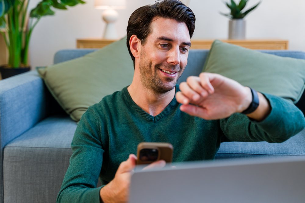 Smiling man wearing a green sweater checks his smartwatch while holding a smartphone seated on a gray couch in a living room with plants, Spain.