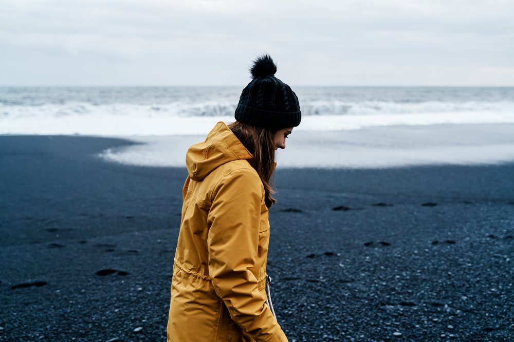 A white woman walks along a black sand beach near the ocean in Iceland