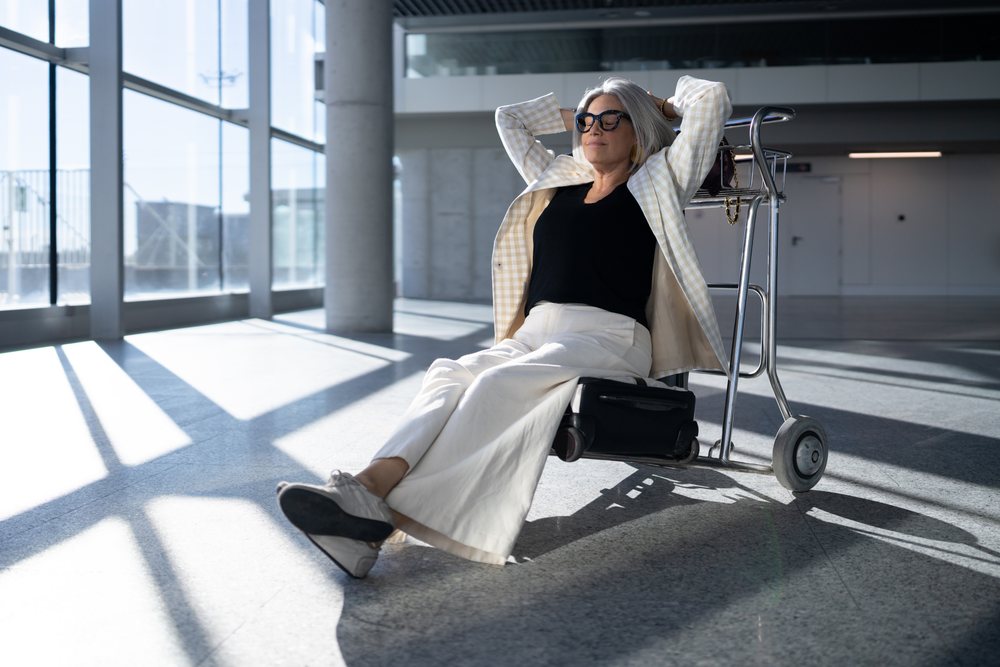 Woman with gray hair and glasses relaxes seated on an airport luggage cart holding a black suitcase in a modern indoor terminal, businesswoman, traveler, Spain, Madrid.