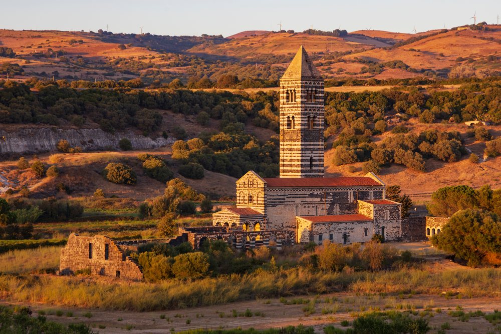 Historic stone Church of the Holy Trinity Saccargia with striped bell tower and ruins in the Sardinian countryside of Codrongianos at sunset, Travel Images, Sardinia, Italy.