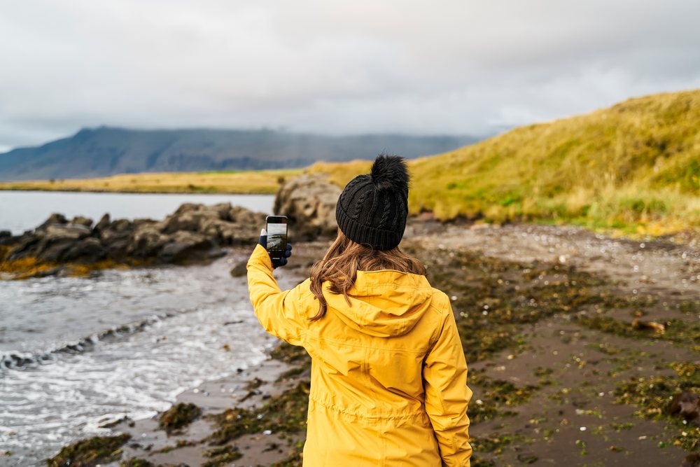 A white woman takes a phone photo on a rocky shore