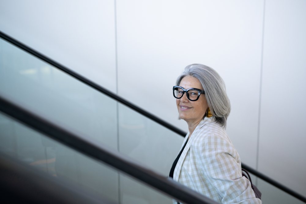 Woman with gray hair wearing eyeglasses and a plaid blazer smiles on an indoor escalator, businesswoman, Spain, Madrid.