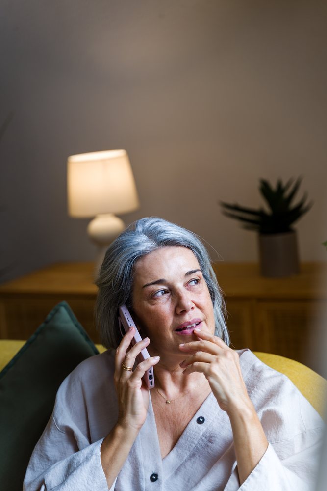 Woman with gray hair sits on a yellow sofa talking on a mobile phone in a living room with a lamp and indoor plant, Spain.