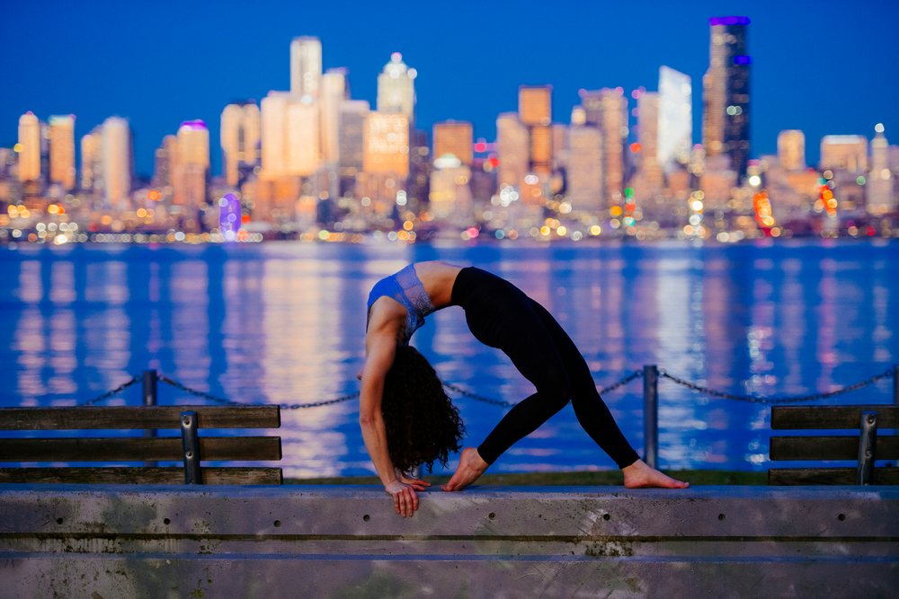 Woman performs a yoga backbend outdoors by the waterfront in Seattle during evening with the city skyline visible, WA, USA.