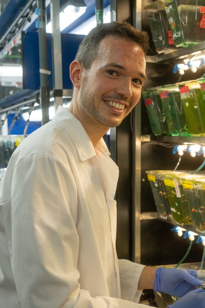A smiling scientist wearing a white lab coat and blue gloves conducts aquatic research in a laboratory, Spain.