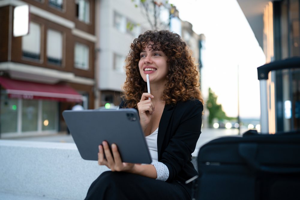 Woman smiling while thinking and holding a tablet with a stylus outdoors in an urban Madrid setting, young business woman.