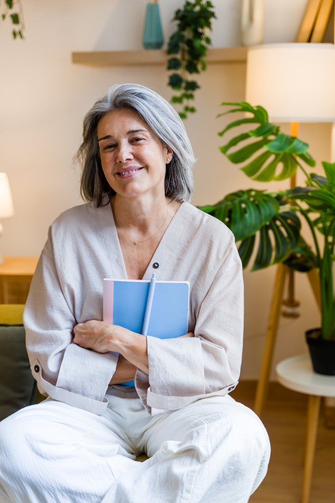 Mature woman sitting indoors smiling and holding a blue notebook beside a monstera plant, Spain.