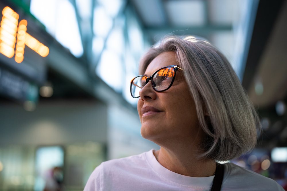 Senior woman with gray hair and reflective eyeglasses wearing a white shirt indoors in a modern public place, Spain.