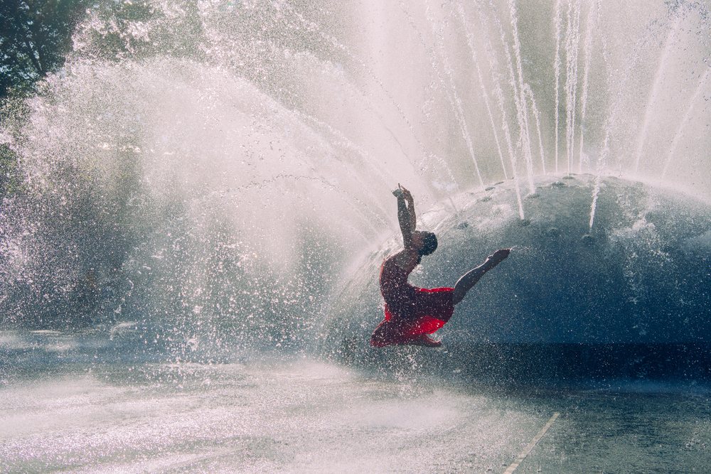 Woman dancer in a red dress leaping near a splashing water fountain outdoors, USA.