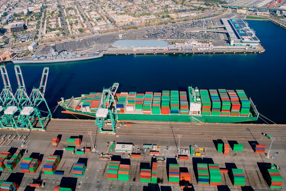 Container ship loaded with colorful shipping containers docked at a major port with cranes and freight yard visible, Port of Long Beach, CA, USA.