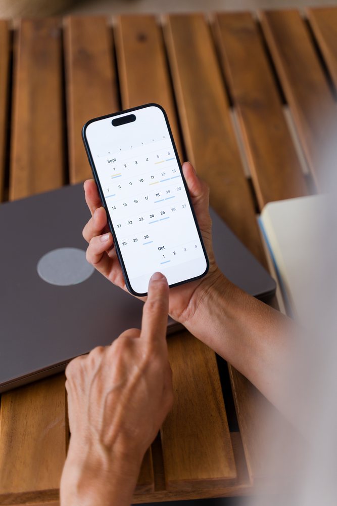 Person holding a smartphone displaying a digital calendar app with scheduled events over a wooden table with a closed laptop, Spain.