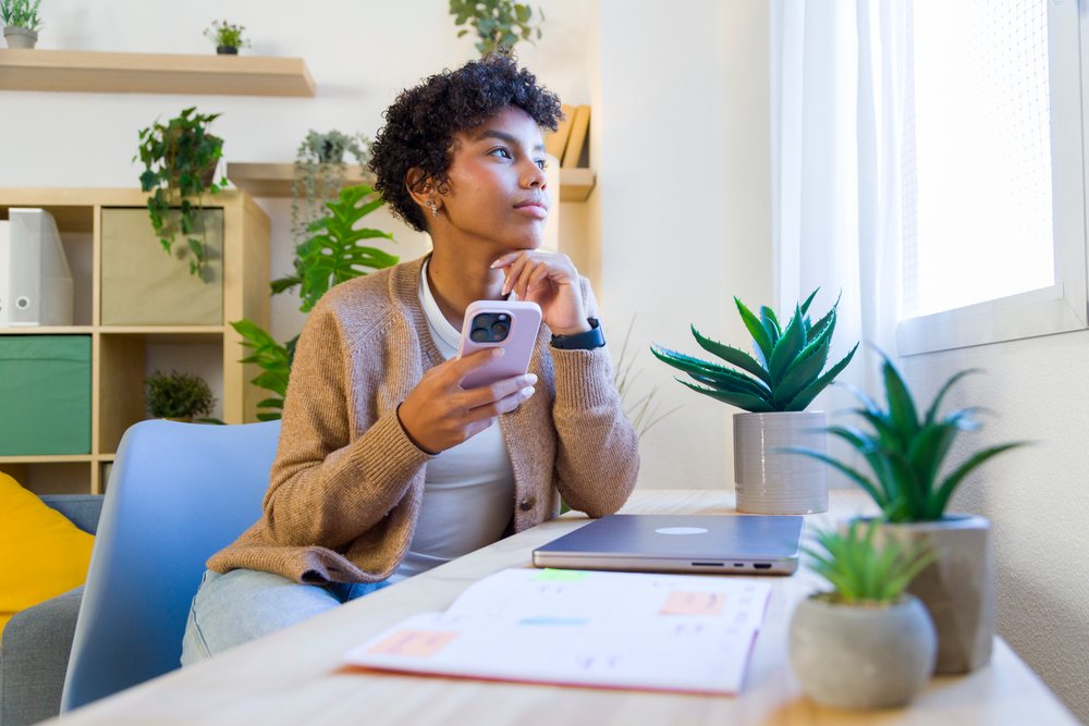 Woman sits at a desk in a modern home office holding a smartphone and looking out a window with daylight illuminating the room, remote work.