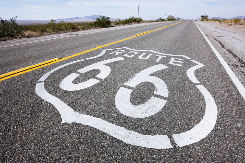 White Route 66 road marking on a two-lane asphalt highway through a desert landscape with bushes and distant mountains, California, USA.