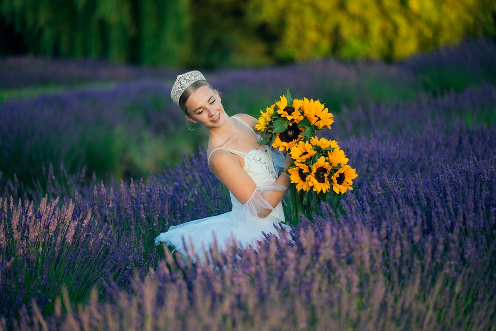 A ballerina wearing a white tutu and tiara smiles while holding a bouquet of sunflowers in a purple lavender field in Washington, WA, USA.