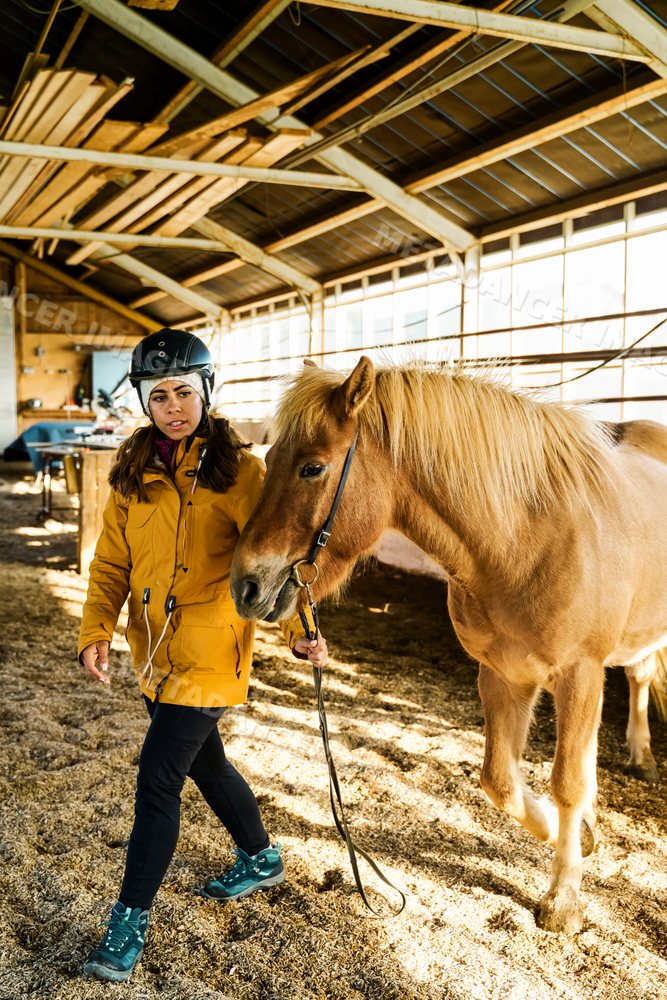 A white woman walks a brown horse in an indoor riding arena