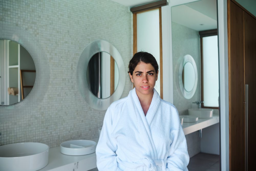 A woman in a white bathrobe stands inside a modern bathroom with circular mirrors on the tiled wall, Maldives.