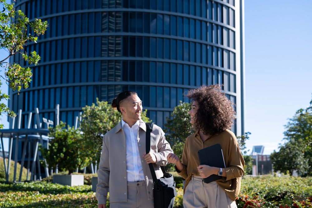 Two business professionals walk and talk in a sunny urban park near a modern office building, Madrid, Spain.