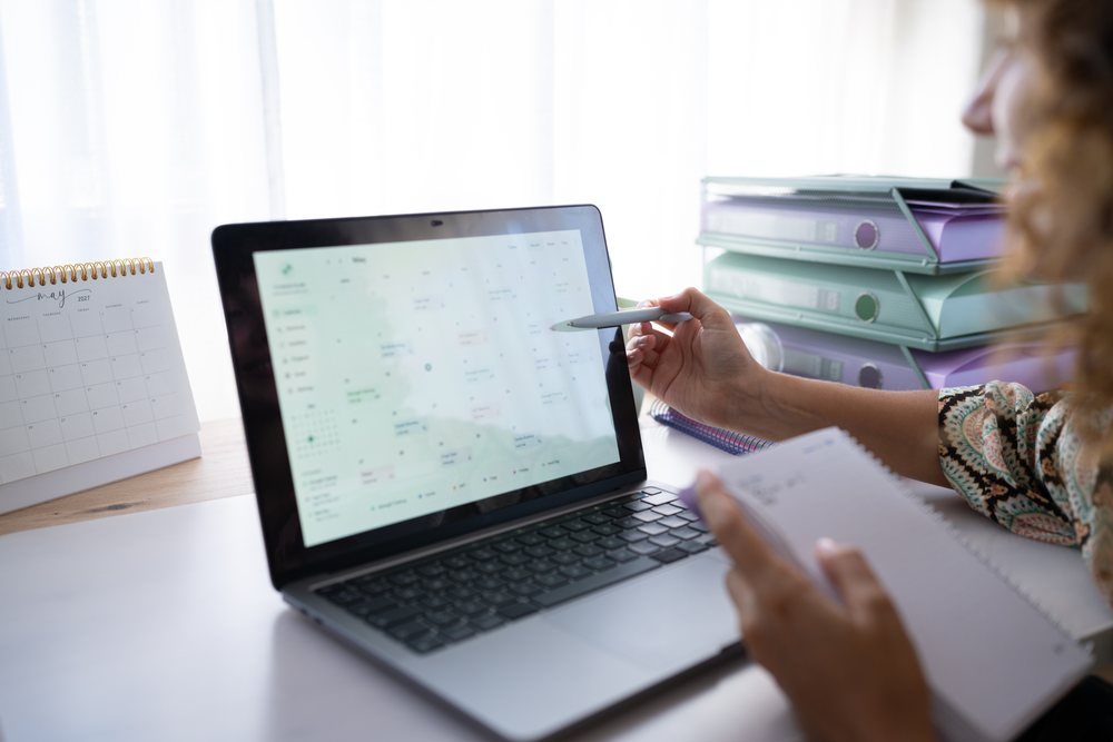 Person planning schedule on laptop with calendar and notebook at desk in a home office setting, Spain.