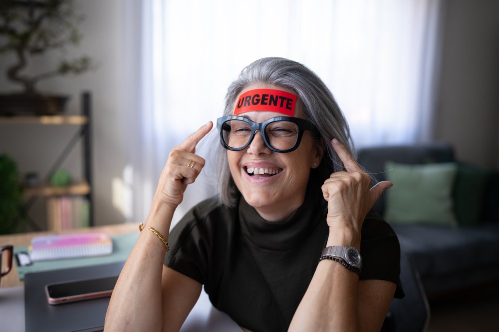Smiling woman with gray hair and glasses points to a red urgente sticker on her forehead in an indoor home office, Spain.