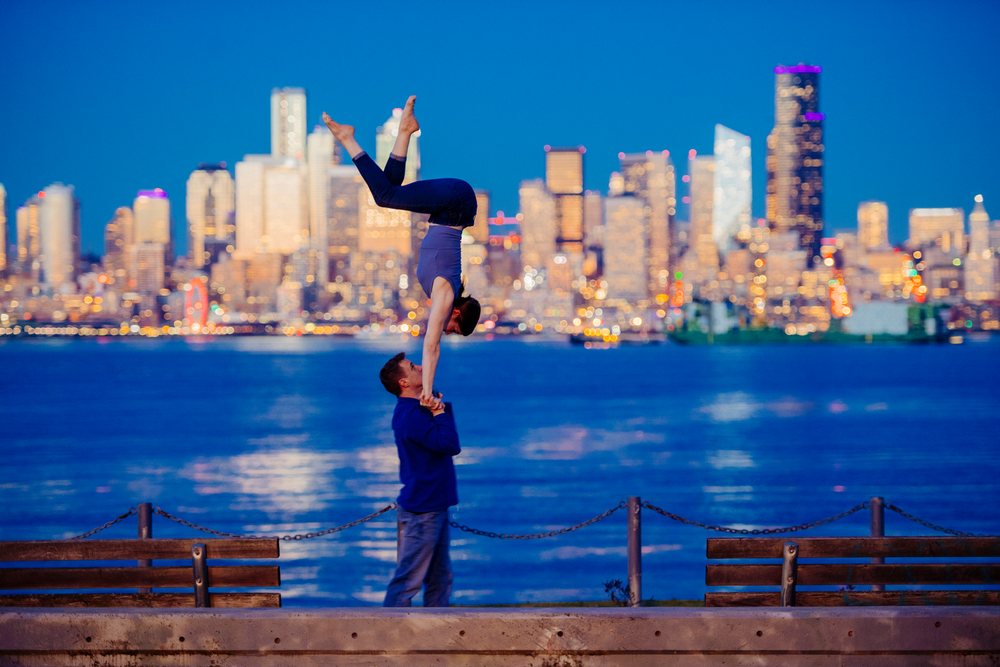 Man holds a woman's hands as she performs a handstand by the Seattle waterfront with the city skyline at dusk, partner acrobatics, yoga, USA.