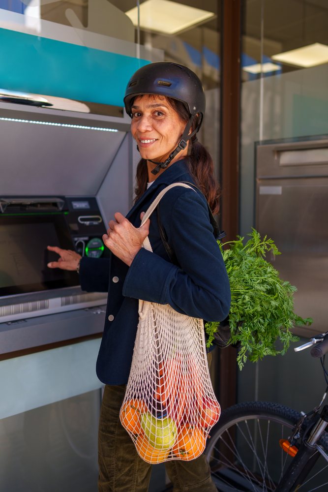 Woman wearing a bicycle helmet uses an ATM while carrying a mesh bag with carrots, oranges, pear, and green leafy vegetable in an urban setting, Málaga.